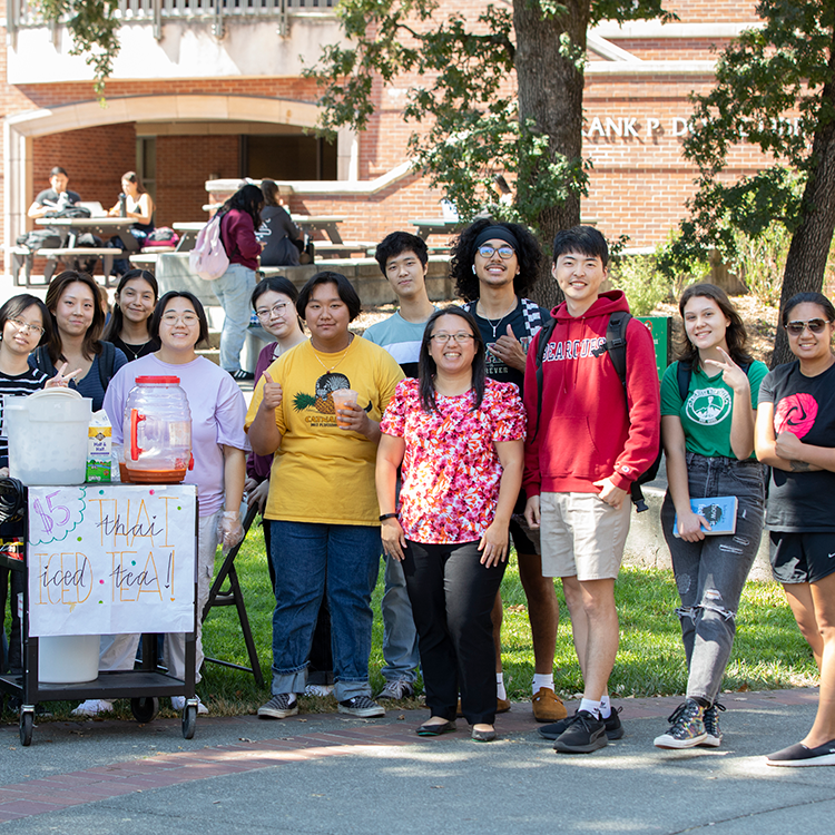A group of Asian American and Pacific Islander students and faculty are posing in front of Doyle Library by a stand where they sell Thai iced tea for a club fundraiser. 